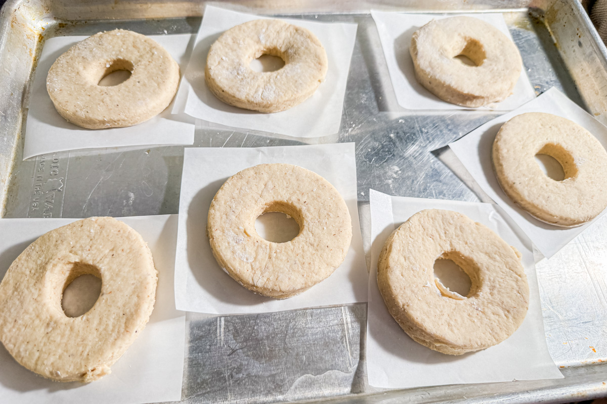 Seven unbaked, ring-shaped dough pieces sit on individual squares of parchment paper, arranged on a metal baking tray, ready to be baked.