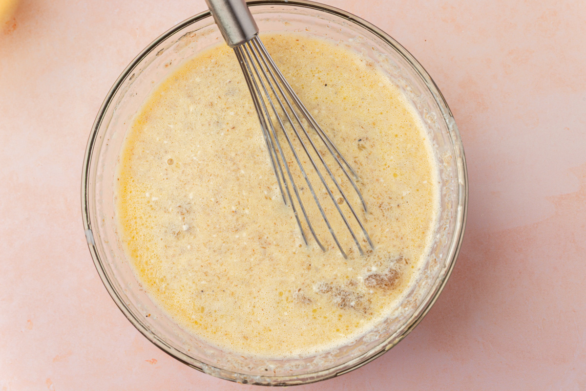 A metal whisk is resting in a glass bowl filled with a light brown, speckled batter mixture on a pale pink surface.