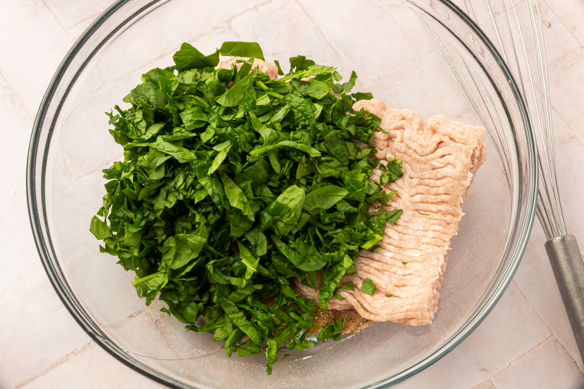 A glass mixing bowl containing raw ground turkey and a pile of chopped fresh spinach, with a metal whisk nearby on a light-colored countertop.