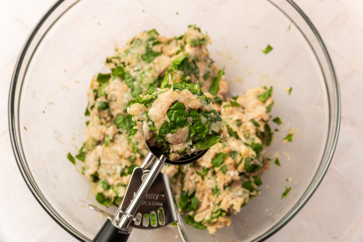A glass bowl filled with a mixture of chopped spinach, coconut flour, ground turkey and other ingredients, with a metal scoop holding a portion of the mixture above the bowl.
