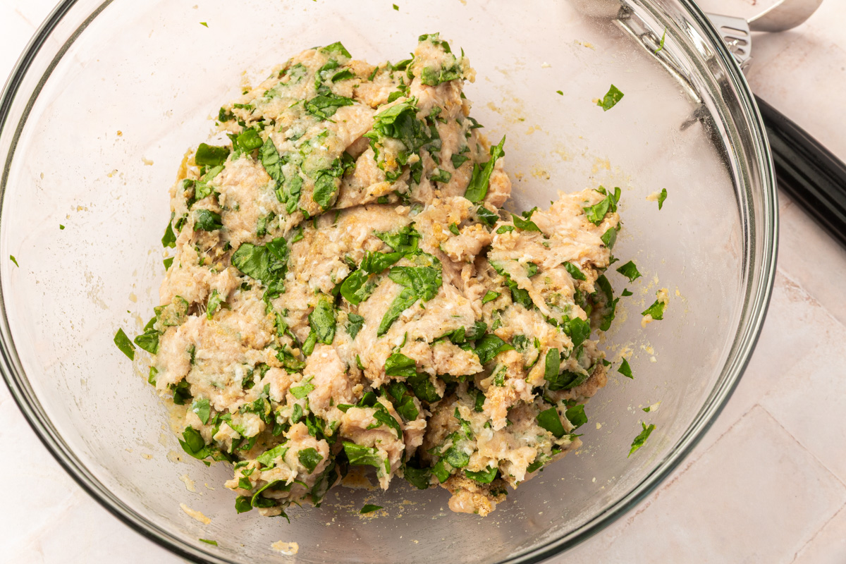 A glass bowl containing a mixture of chopped green herbs, coconut flour, and ground turkey sits on a warm white countertop. A metal utensil rests nearby.
