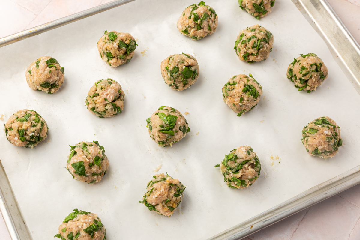 Uncooked meatballs mixed with chopped green herbs are arranged in rows on a parchment-lined baking sheet, ready to be cooked.
