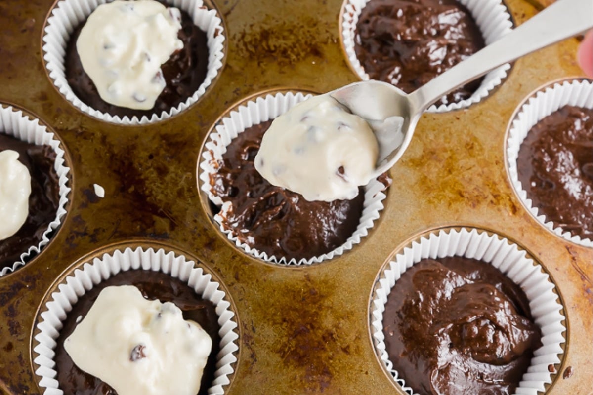 A close-up of a muffin tin filled with paper liners. Some liners contain chocolate batter, while others have chocolate batter topped with a spoonful of white batter being added.