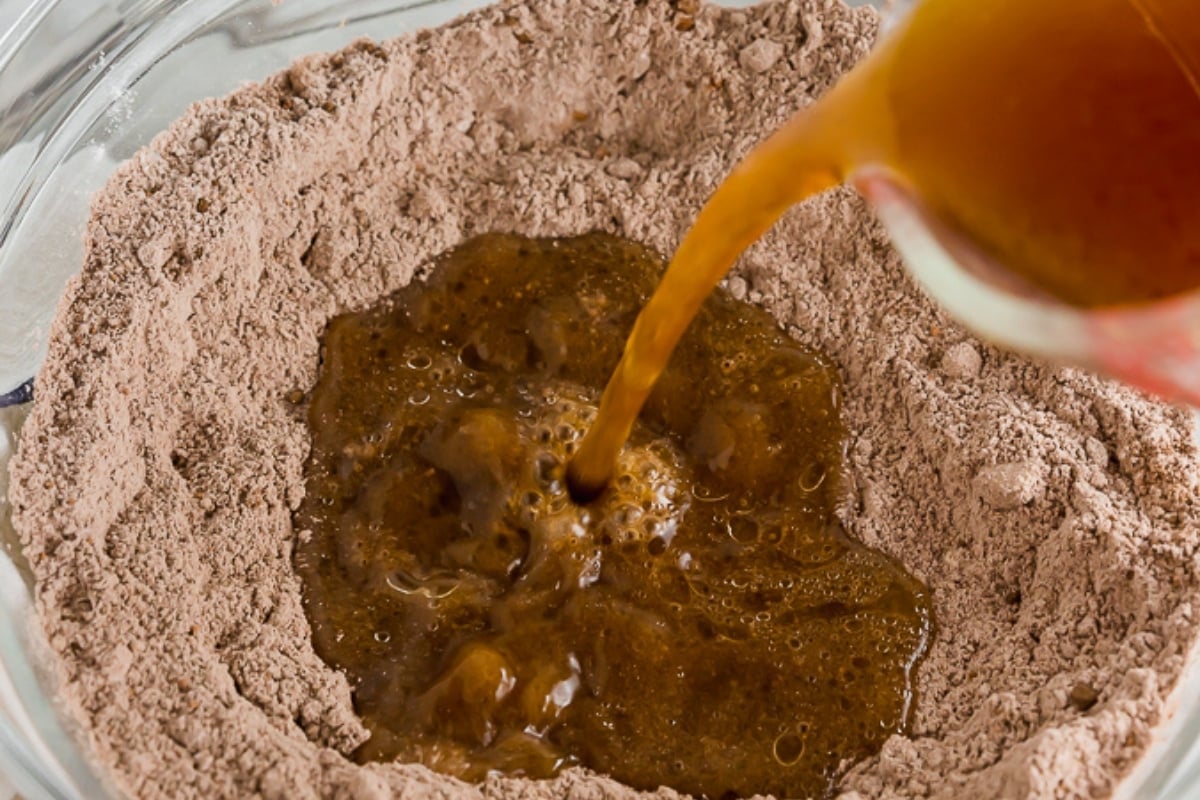 A glass bowl with dry chocolate cake mix has a well in the center, where brown liquid is being poured in from a glass measuring cup.
