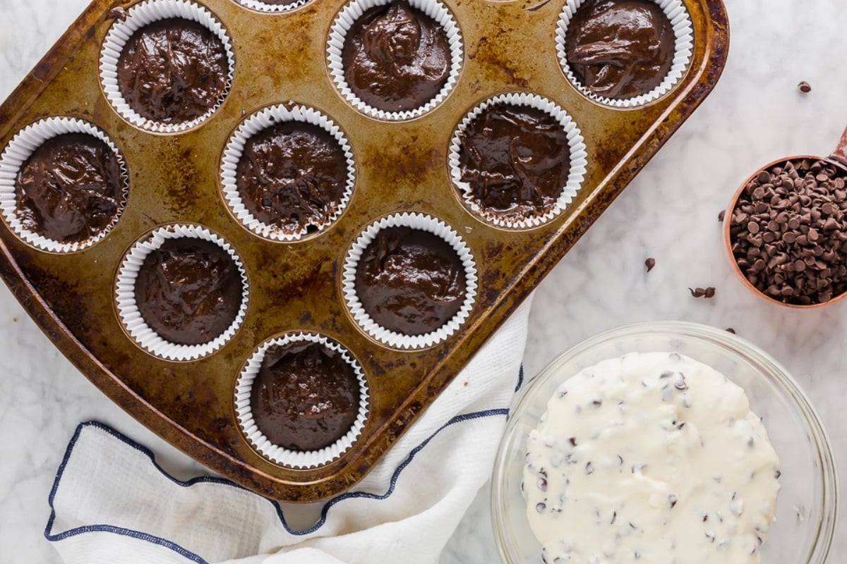 A muffin tin filled with cupcake liners, each holding chocolate batter, sits on a marble surface next to a bowl of white batter with chocolate chips and a copper cup filled with mini chocolate chips.