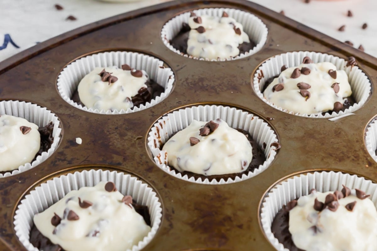 A muffin tin holds several paper liners filled with chocolate batter and topped with cream cheese mixture and chocolate chips, ready to be baked.