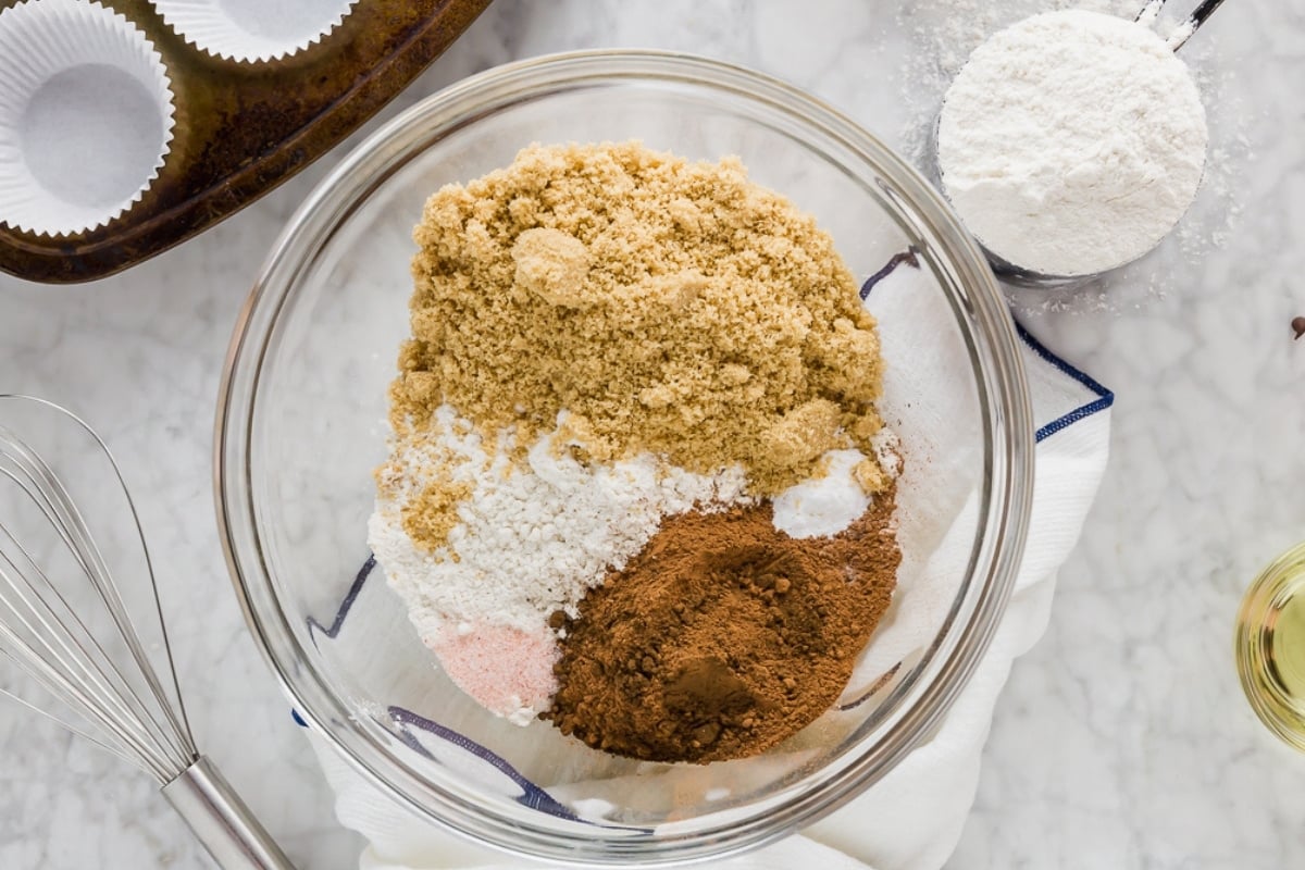 A glass bowl filled with brown sugar, flour, baking powder, and cocoa powder sits on a white towel, surrounded by cupcake liners, a whisk, a measuring cup of flour, and a small container of oil.