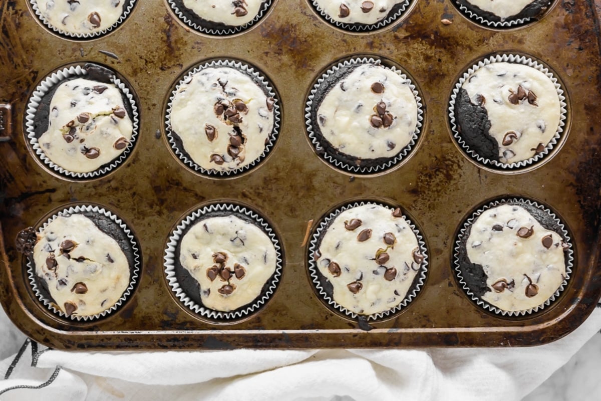 A muffin tin filled with chocolate chip muffins in paper liners, some still unbaked, on a worn baking tray with a white cloth underneath.