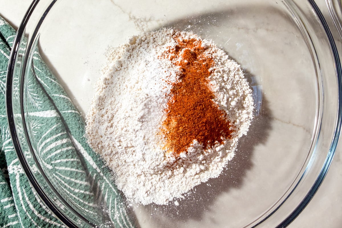 A glass bowl on a marble surface contains flour and reddish spices, with a green and white patterned kitchen towel beside it.