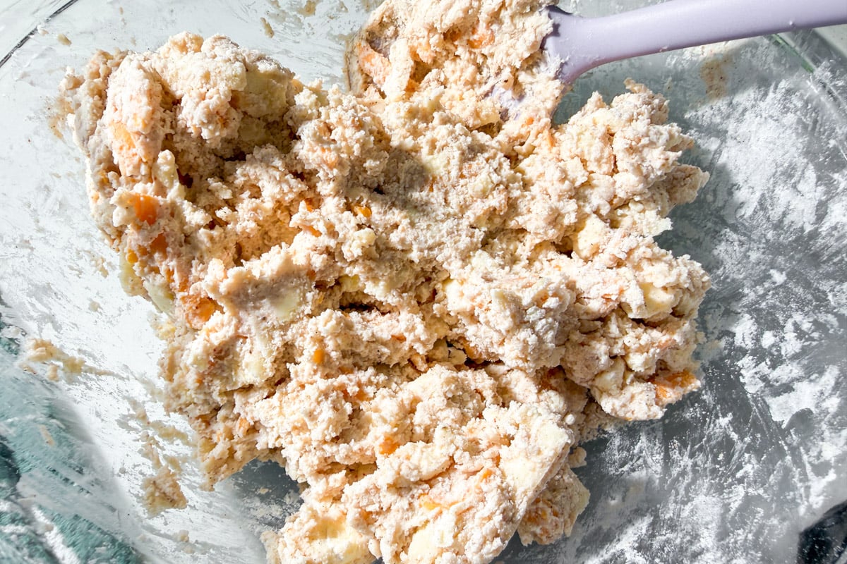 A glass bowl containing crumbly biscuit or dough mixture, with visible chunks of butter and flour, being mixed with a purple spatula.