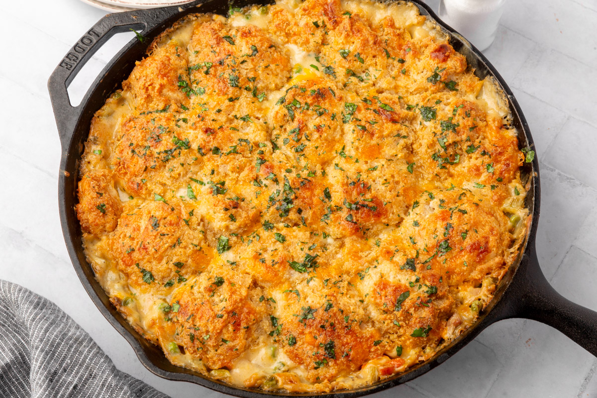 A cast iron skillet filled with a golden-brown, baked casserole topped with biscuit rounds and sprinkled with fresh herbs, sitting on a light countertop next to a striped cloth.