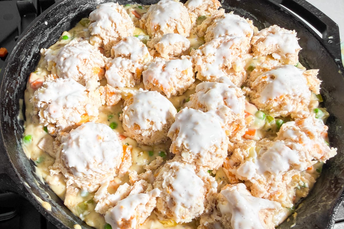 A skillet filled with creamy chicken and vegetable mixture, topped with unbaked biscuit dough portions, ready to be baked.