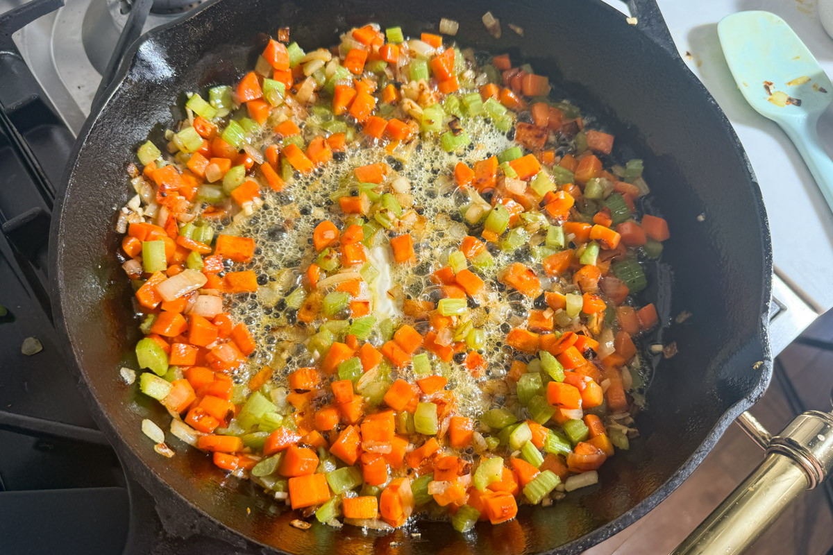 Diced carrots, celery, and onions are sautéing in melted butter in a black cast iron skillet on a stove.