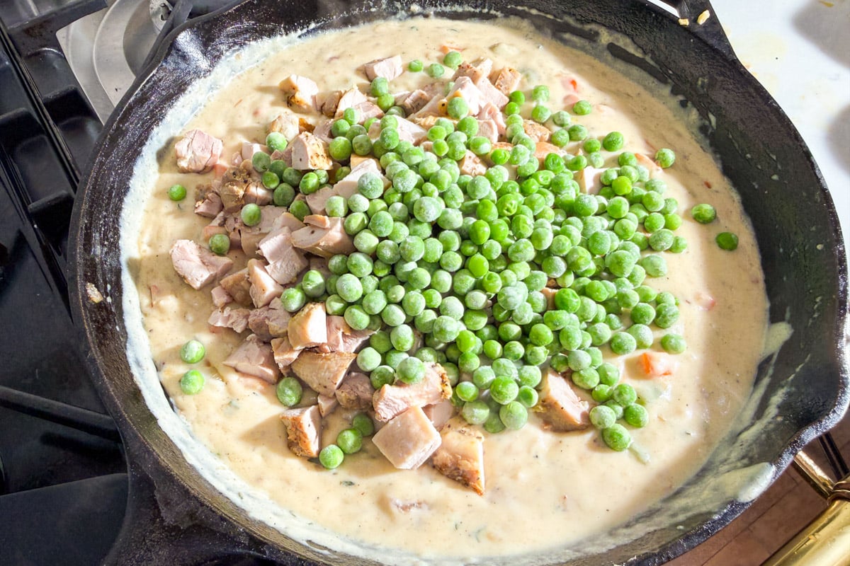 A skillet filled with creamy sauce, diced cooked chicken, and a mound of frozen green peas on top, being prepared on a stovetop.