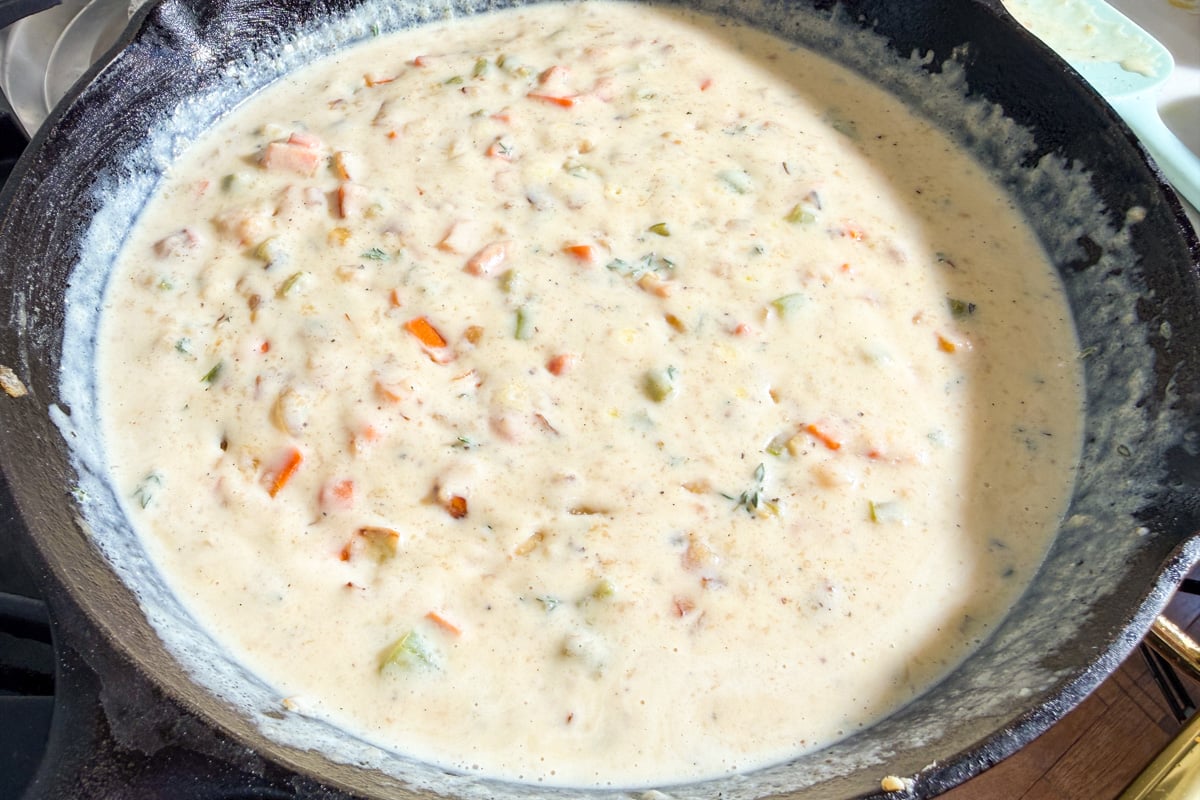 A close-up of a skillet filled with a creamy white sauce containing chopped vegetables such as carrots, celery, and possibly onions and herbs. The mixture is simmering on a stovetop.
