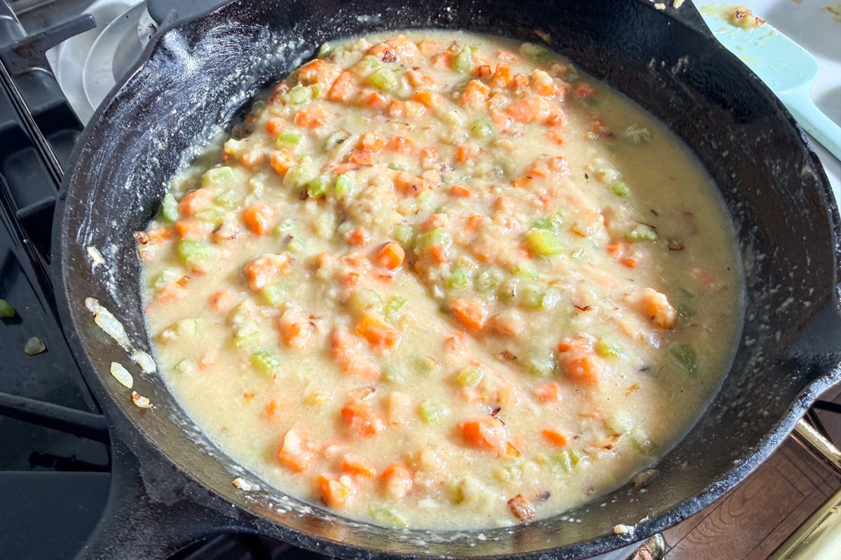 A cast iron skillet on a stove contains a creamy mixture of chopped carrots and celery being cooked with chicken broth for a chicken pot pie.