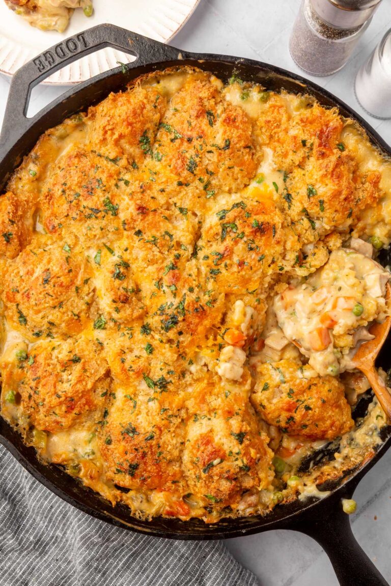A cast iron skillet filled with golden-brown, biscuit-topped chicken pot pie, showing creamy filling with vegetables like carrots and peas, and a serving being scooped out with a wooden spoon.
