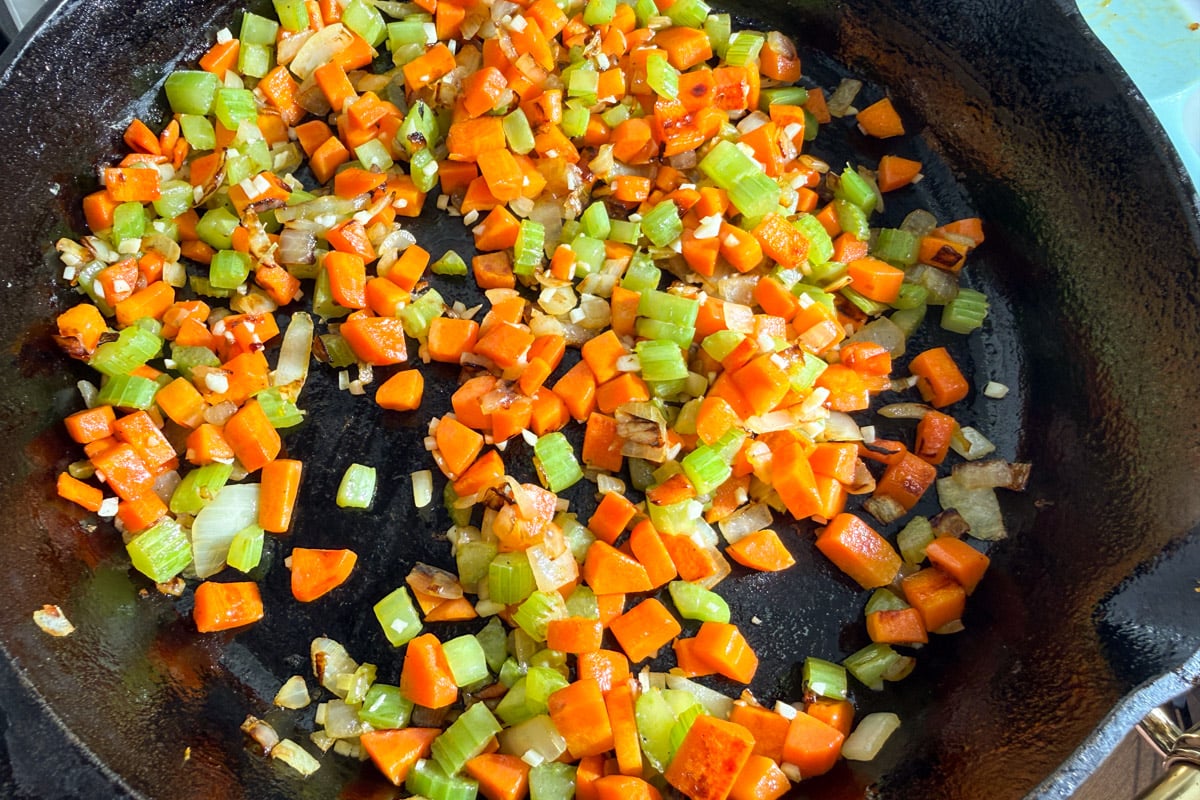 A cast iron skillet filled with diced carrots, celery, and onions being sautéed, with vegetables lightly browned and glistening.