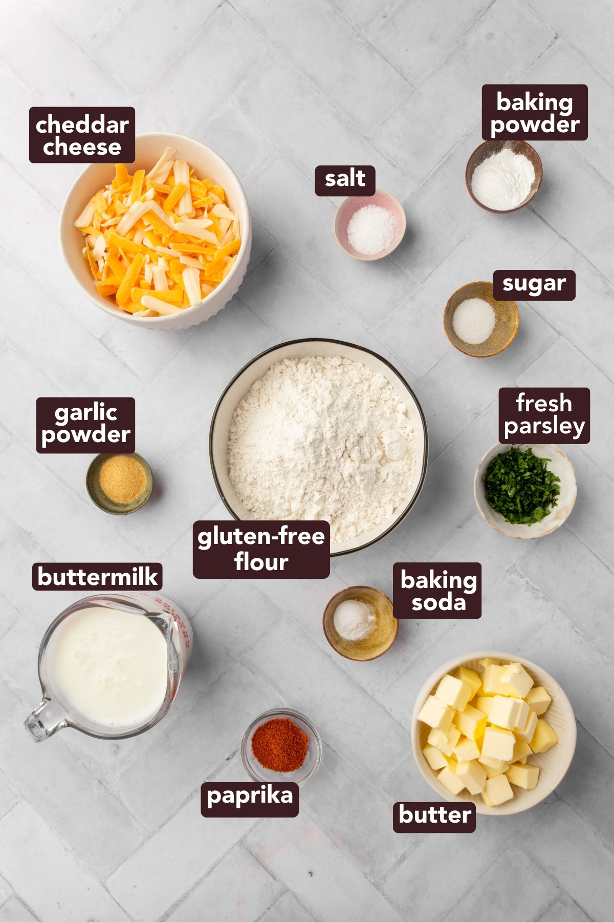 A flat lay of labeled ingredients for gluten-free cheddar biscuits, including cheddar cheese, salt, baking powder, sugar, fresh parsley, gluten-free flour, baking soda, butter, paprika, buttermilk, and garlic powder on a white surface.