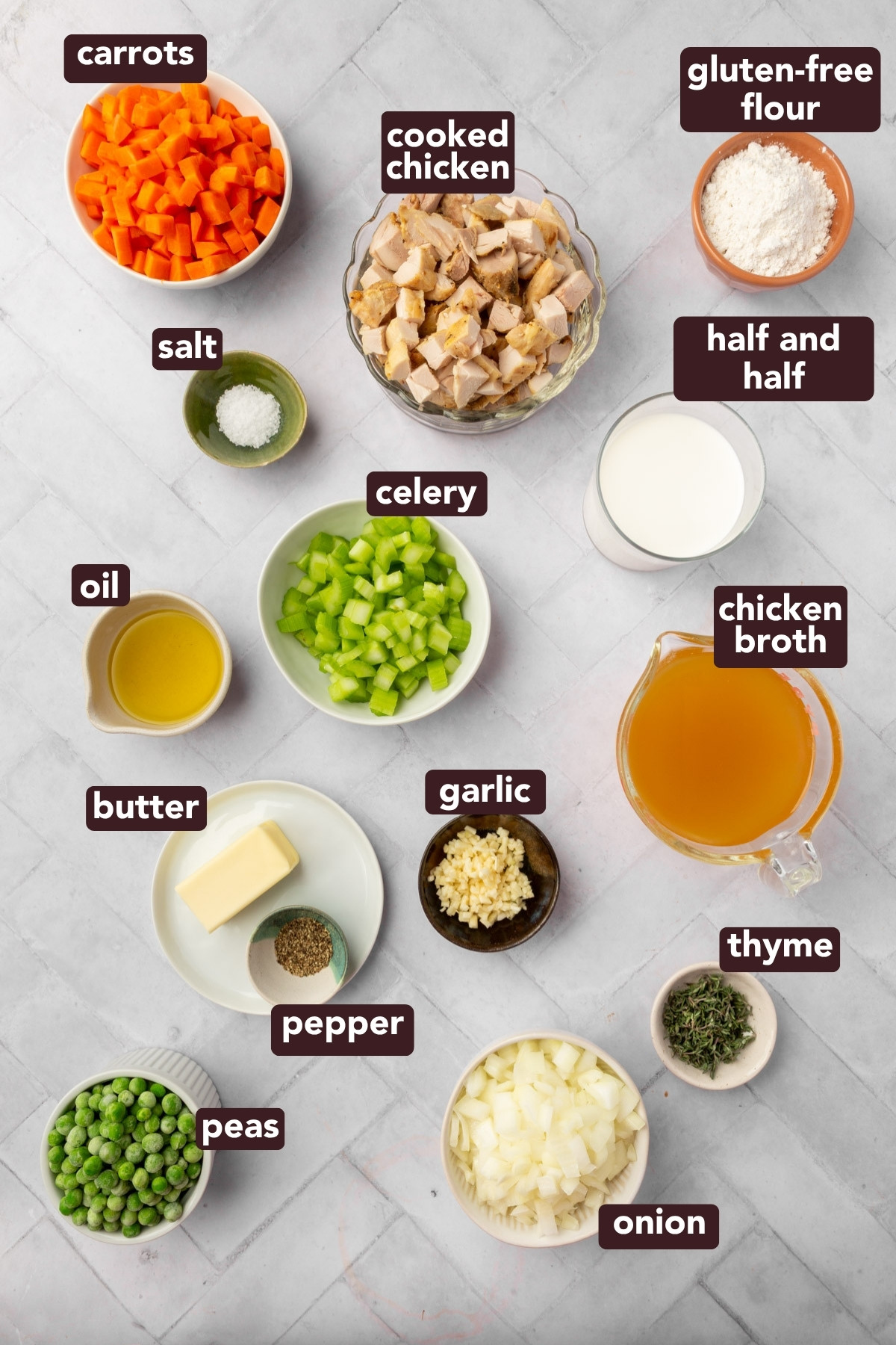 Top-down view of various labeled ingredients in bowls on a white surface, including carrots, cooked chicken, gluten-free flour, half and half, chicken broth, celery, salt, oil, butter, garlic, thyme, onion, pepper, and peas.