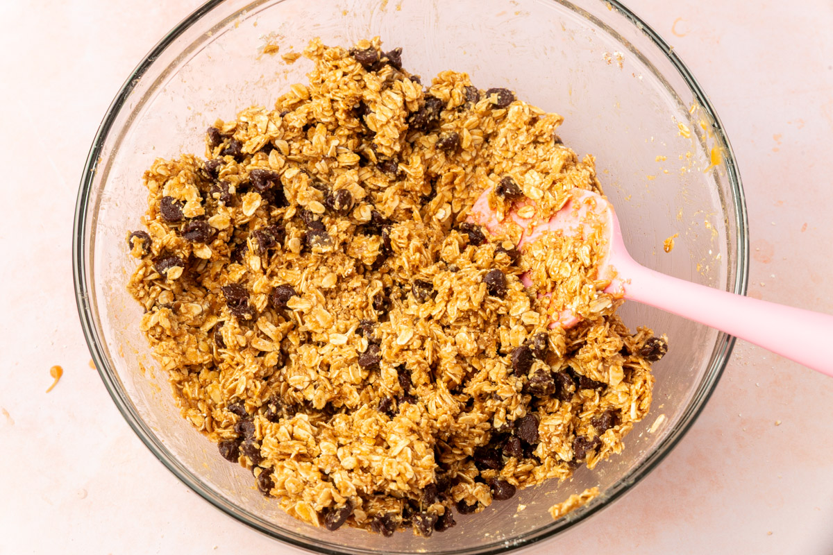 A glass bowl filled with a mixture of oats, raisins, and a sticky batter. A pink spatula is partially submerged, mixing the ingredients together on a light-colored surface.