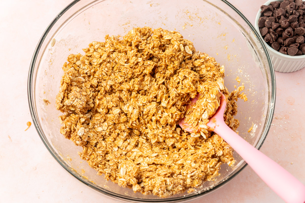 A glass bowl filled with a mixture of oats and a light brown batter, being stirred with a pink spatula. Next to the bowl is a small white dish of chocolate chips on a light pink surface.