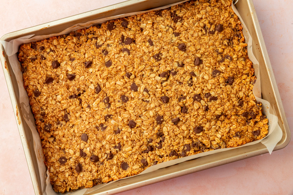 A rectangular baking pan lined with parchment paper holds a large, baked oat bar with visible chocolate chips and a golden-brown color.