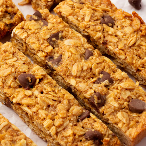 Homemade gluten-free peanut butter granola bars with chocolate chips are arranged on parchment paper. The bars have a golden-brown color and visible oats, with some chocolate chips scattered throughout and nearby.