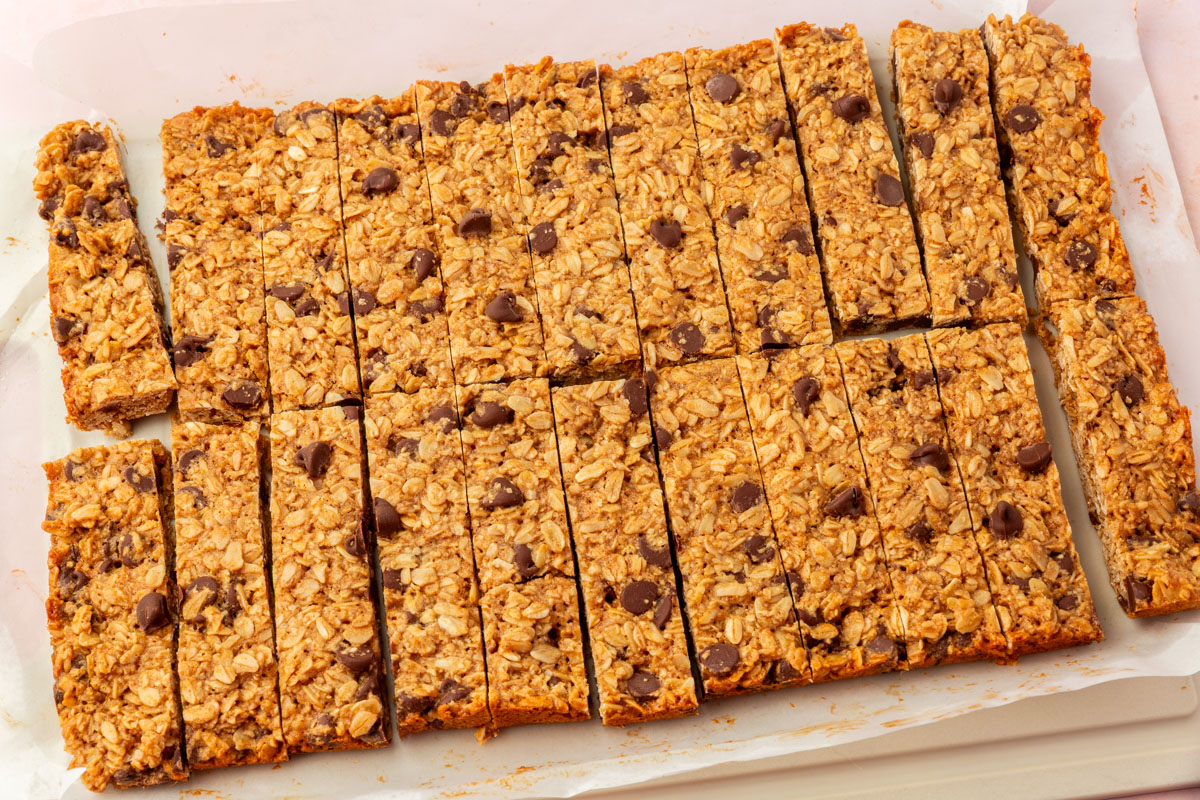 A tray of homemade granola bars cut into rectangular pieces, with visible oats and scattered chocolate chips on top, resting on parchment paper.