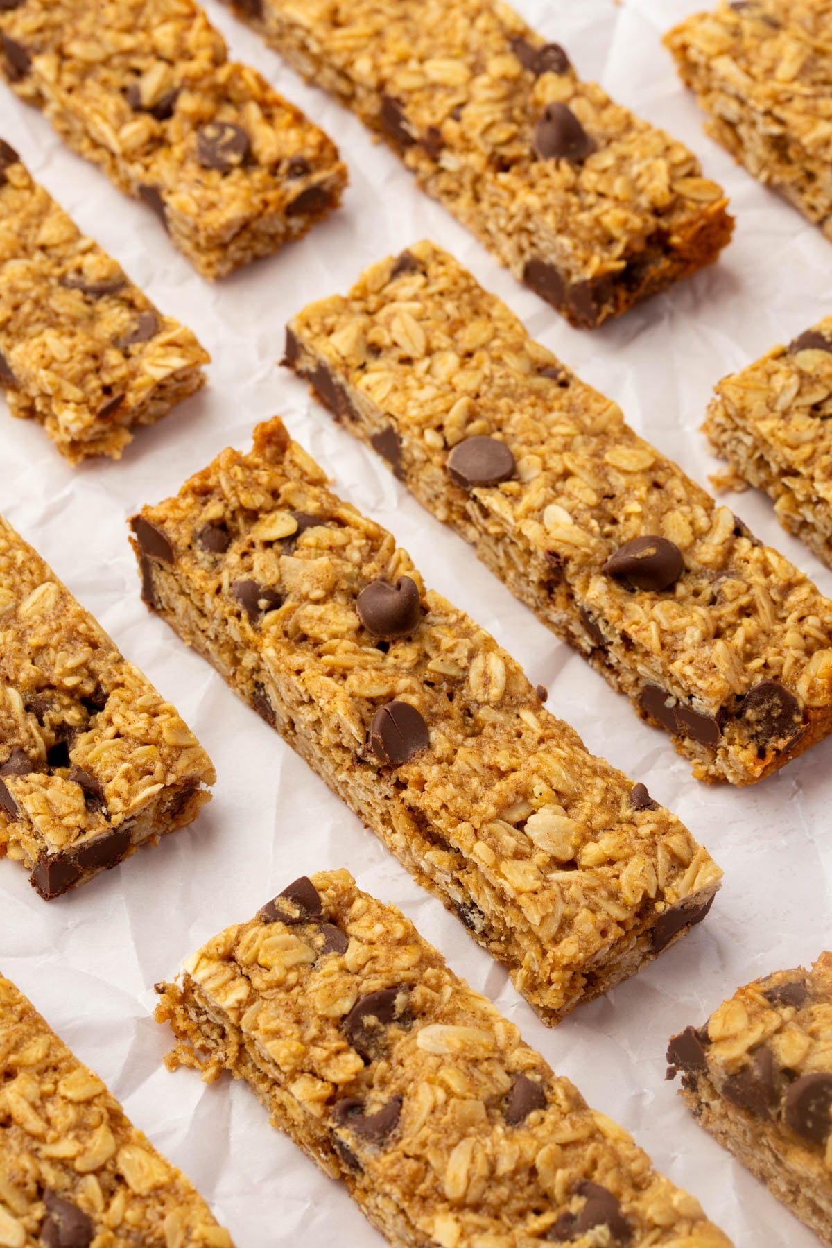 Rows of homemade gluten-free granola bars with gluten-free oats and chocolate chips are arranged on crinkled white parchment paper. The bars are rectangular and appear chewy with visible chocolate pieces.