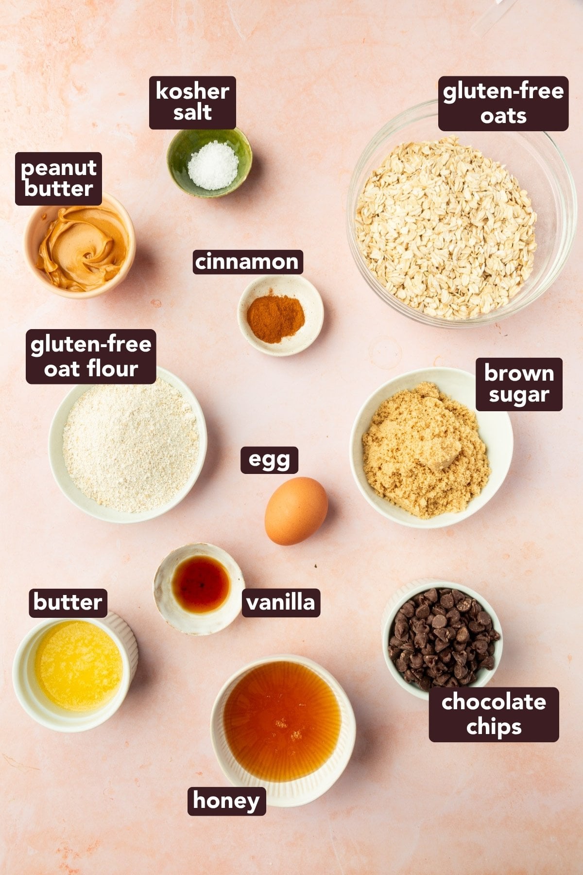 Top-down view of labeled ingredients in bowls on a light surface, including gluten-free oats, gluten-free oat flour, peanut butter, butter, egg, honey, brown sugar, chocolate chips, vanilla, cinnamon, and kosher salt.