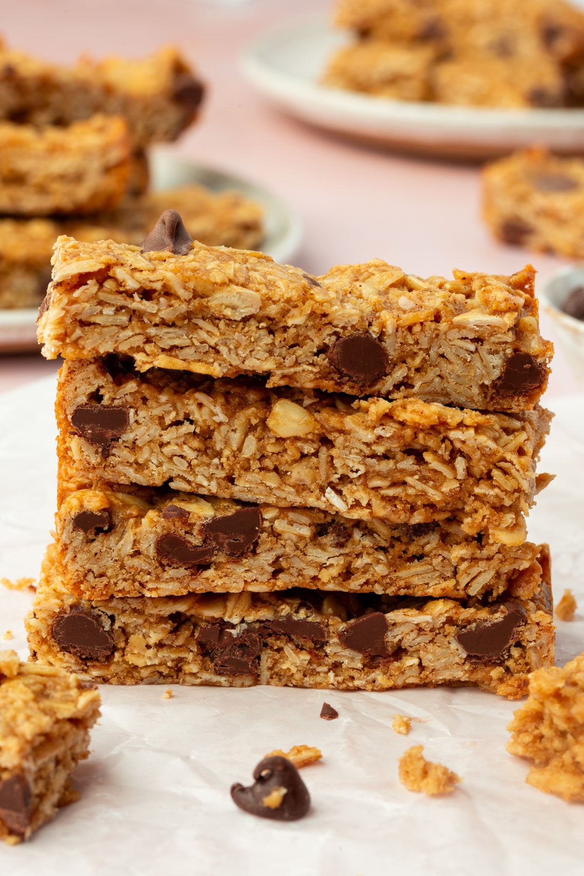 A stack of three homemade gluten-free oatmeal bars with chocolate chips sits on parchment paper, with more bars blurred in the background on plates.