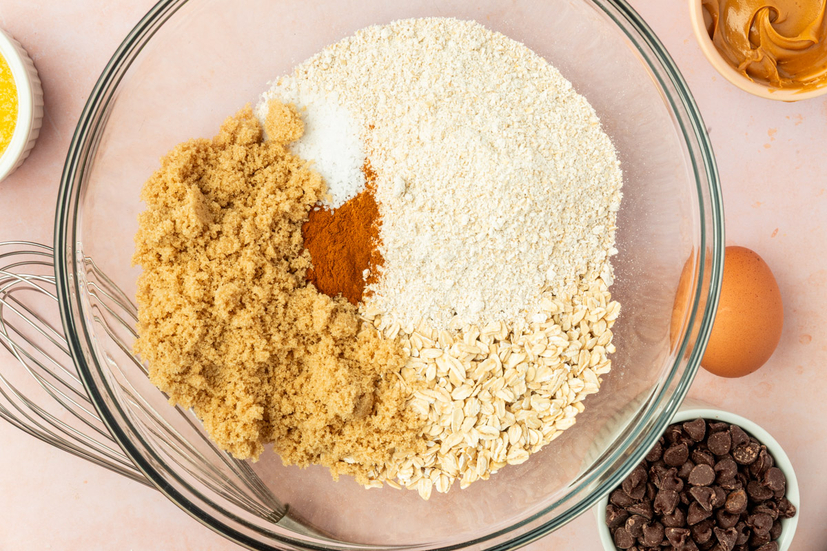 A large glass bowl contains brown sugar, gluten-free oats, gluten-free oat flour, baking soda, salt, and cinnamon, ready to be mixed. Nearby are a whisk, a bowl of chocolate chips, a whole egg, and other ingredients.