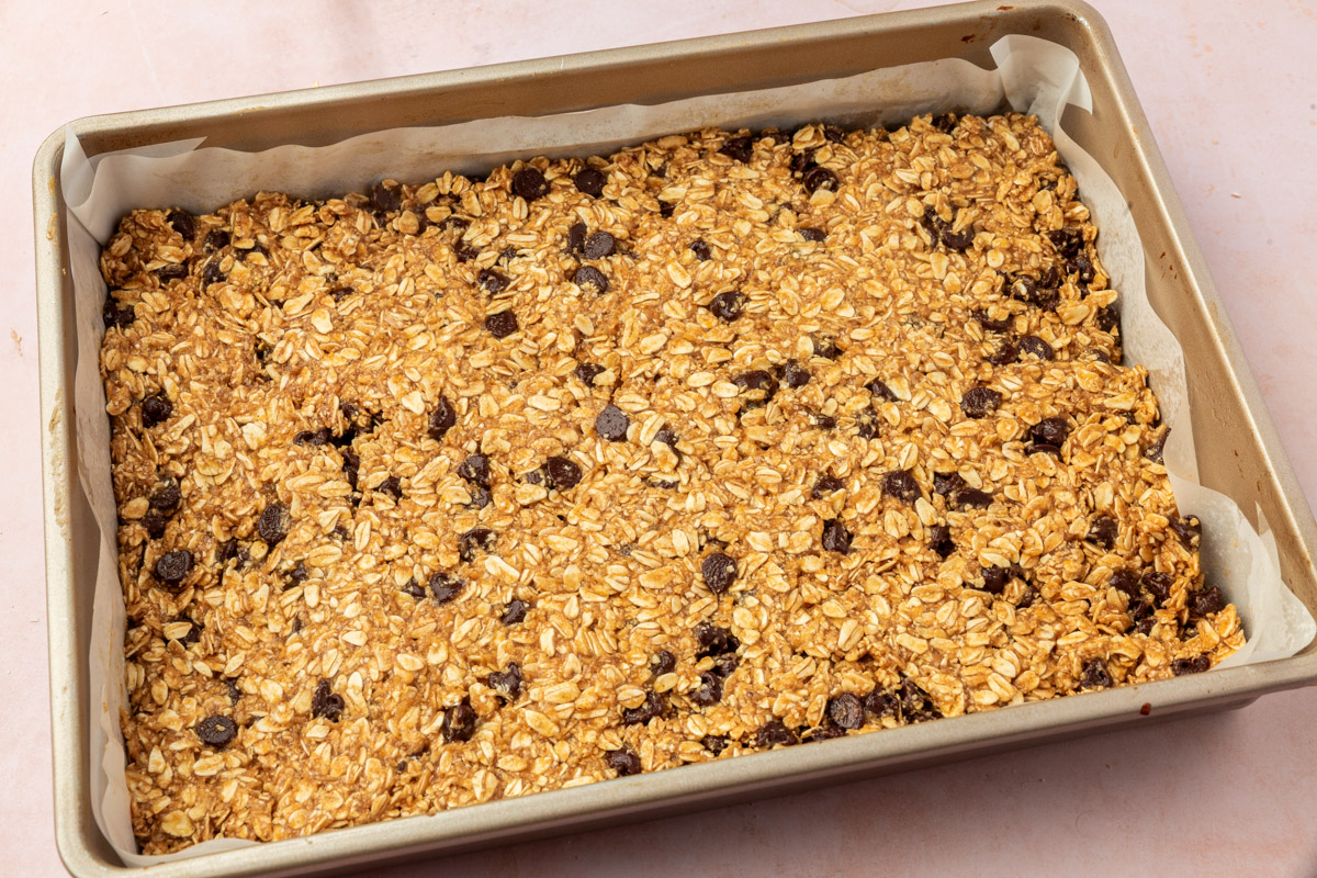 A rectangular pan lined with parchment paper is filled with an unbaked mixture of oats and raisins, ready to be made into granola bars.