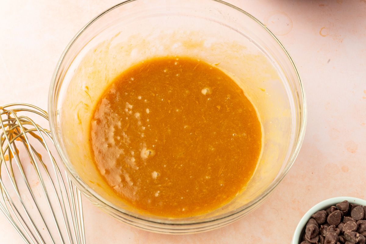 A glass bowl filled with a smooth, orange-brown batter sits on a light surface. A metal whisk is next to the bowl, and a small bowl of chocolate chips is partially visible nearby.