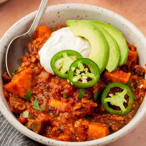 A bowl of chili topped with avocado slices, jalapeño rings, and a dollop of sour cream, served with a spoon. The chili contains chunks of vegetables and is garnished with chopped herbs.