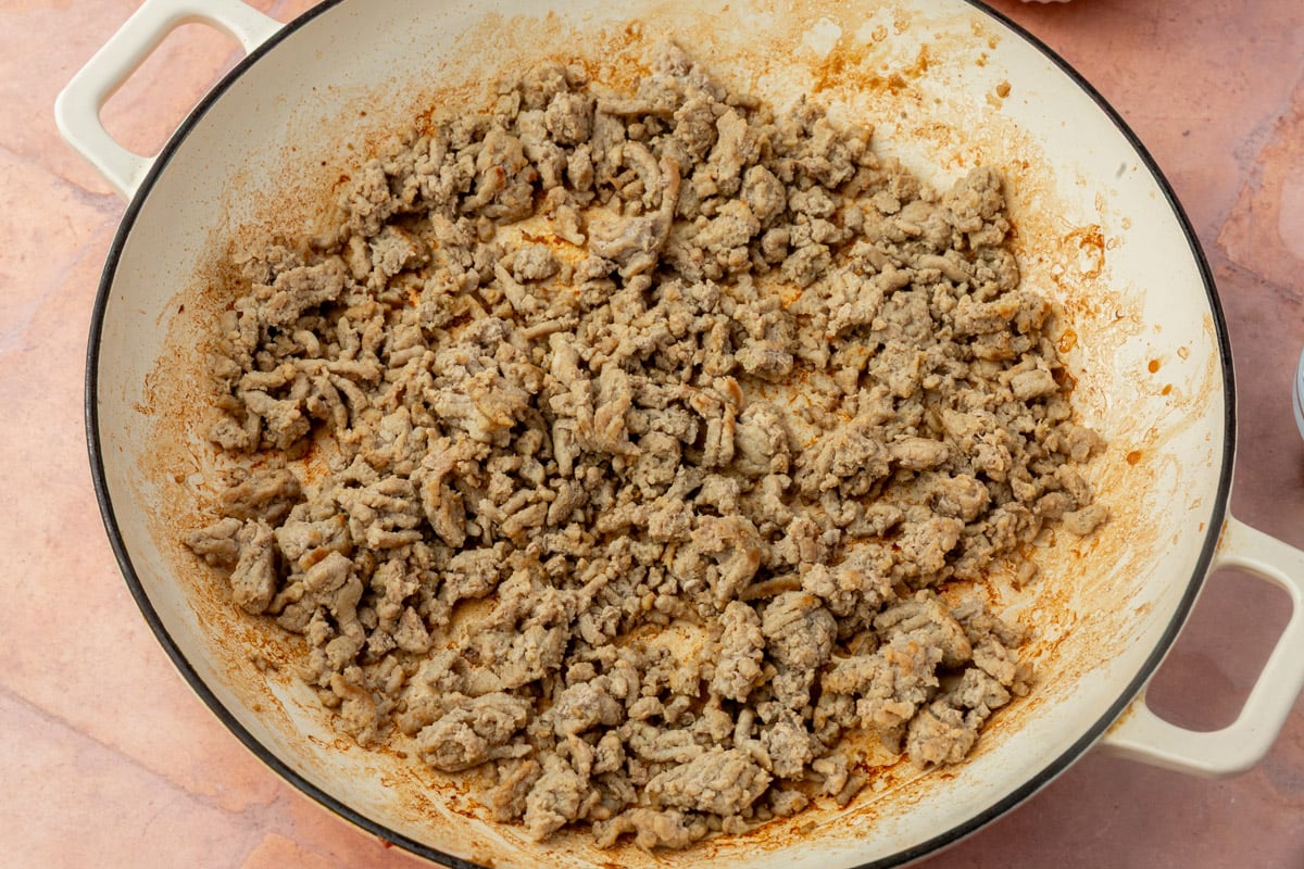 Cooked and seasoned ground turkey is spread out in a large white skillet on a pinkish countertop. The meat is crumbly and lightly browned, with some oil and seasoning residue around the edges of the pan.