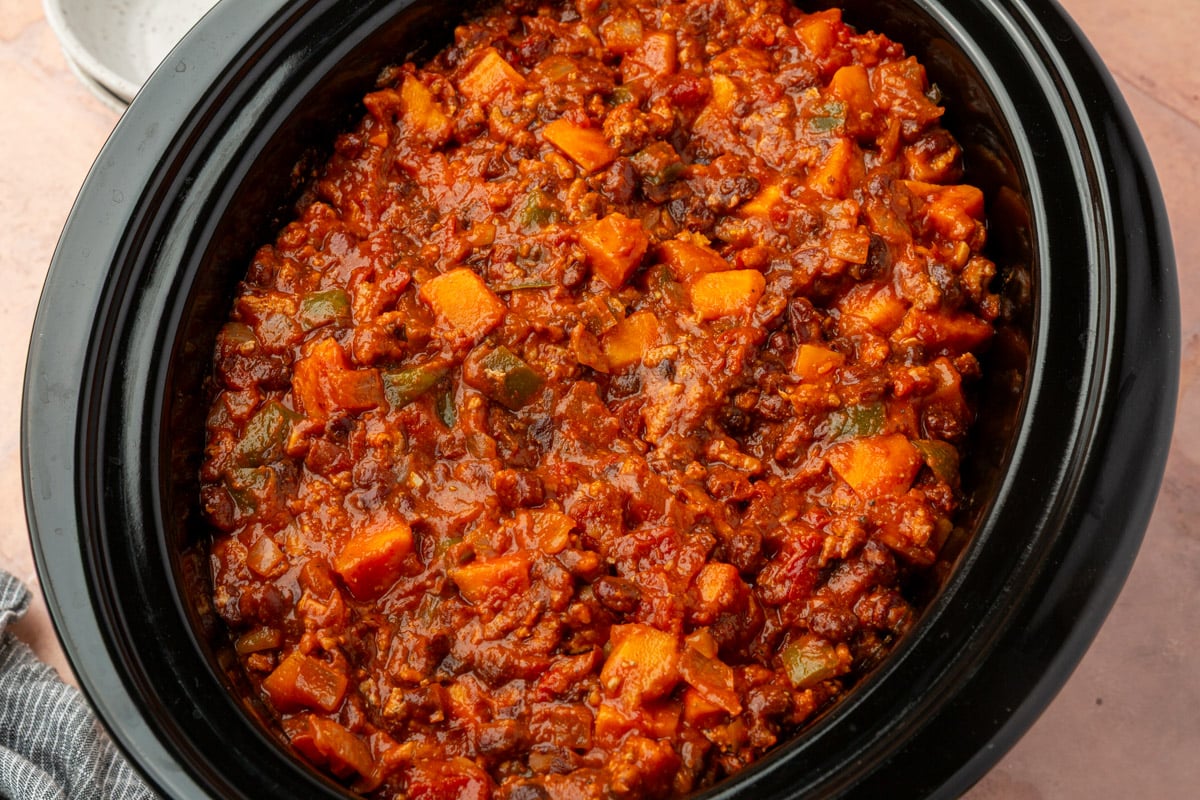 A close-up of a slow cooker filled with chunky vegetable chili, featuring diced carrots, bell peppers, beans, and a rich tomato-based sauce.