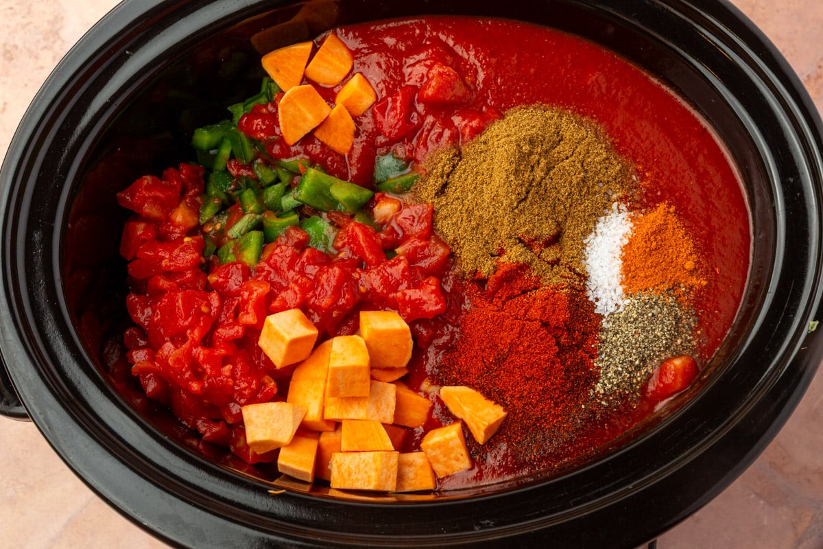 A slow cooker filled with chopped sweet potatoes, diced tomatoes, green bell peppers, tomato sauce, and piles of spices including cumin, chili powder, salt, pepper, and paprika, all before mixing.