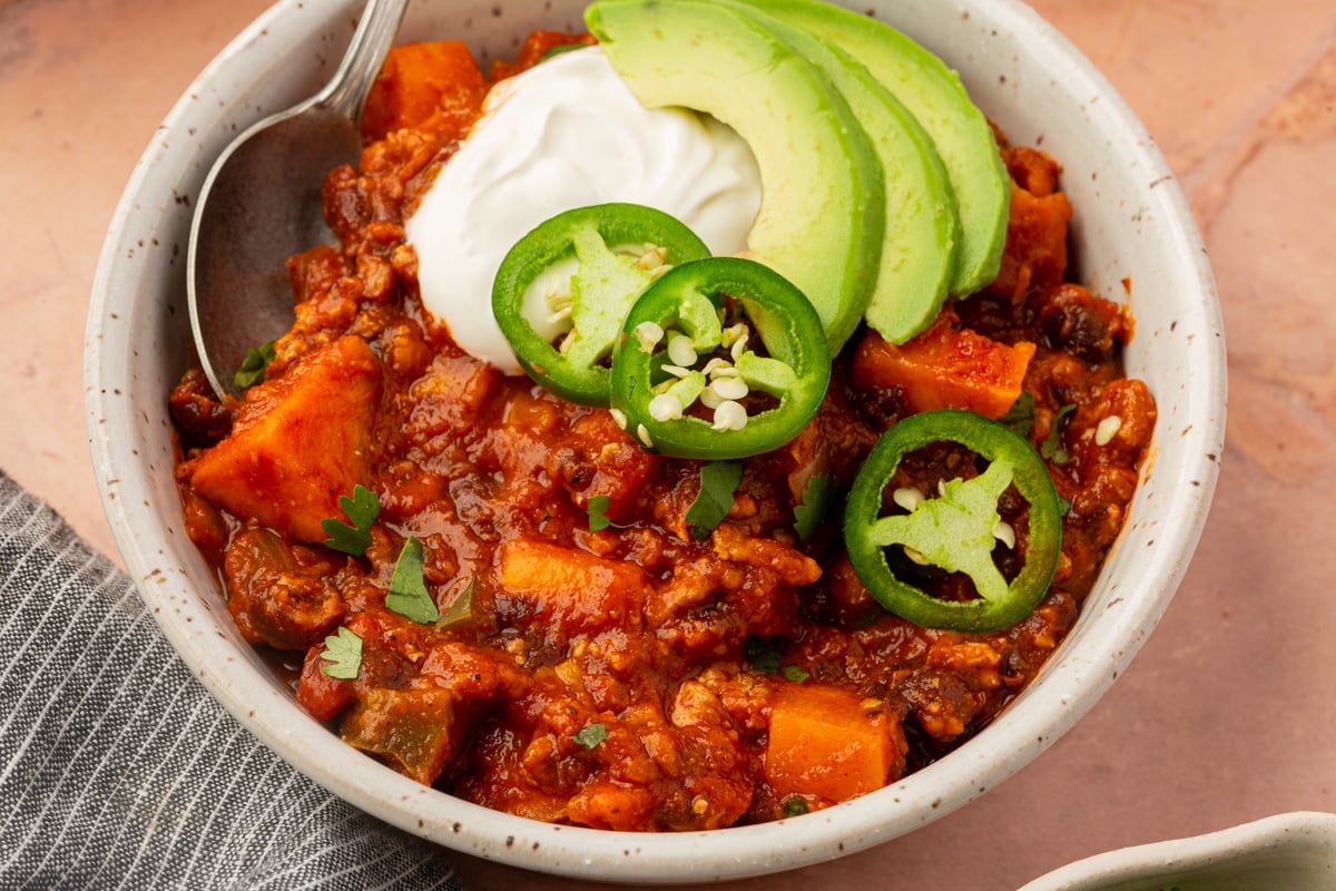 A bowl of chili topped with sliced avocado, jalapeños, and a dollop of sour cream, with a spoon on the side and a gray napkin partially visible.