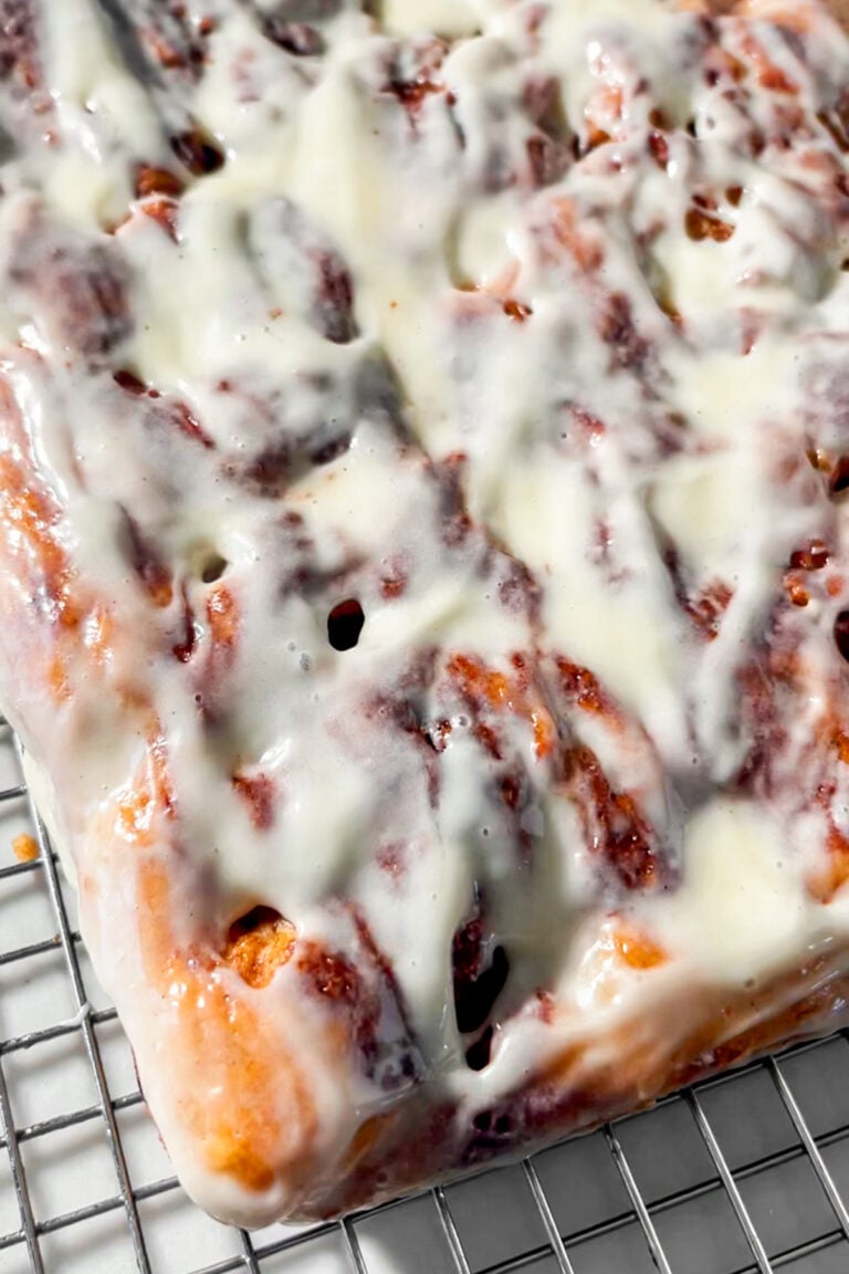 A close-up of a rectangular gluten-free cinnamon roll focaccia slab covered in creamy white icing, resting on a wire cooling rack. The icing is slightly melted and glossy over the swirled, golden-brown pastry.