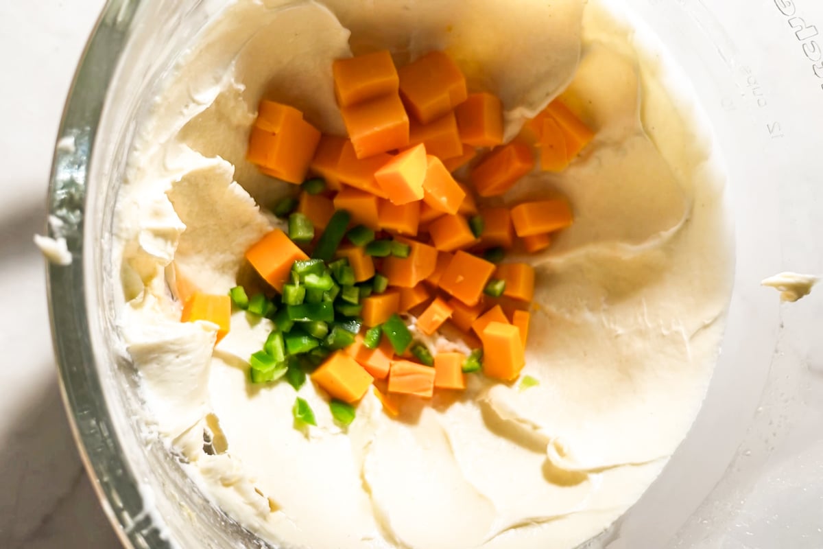 A glass bowl containing creamy focaccia dough with cubed cheddar cheese and chopped jalapeños on top, ready to be mixed.