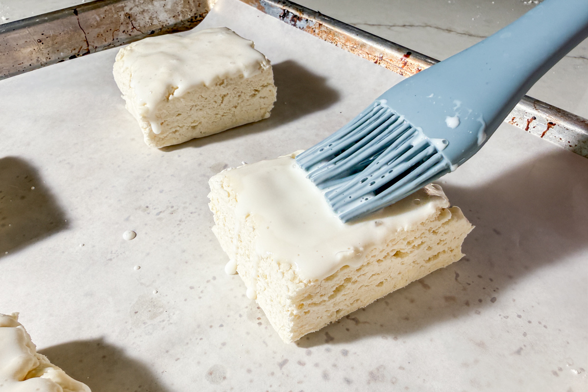 A close-up of an unbaked biscuit on a parchment-lined baking sheet, being brushed with a creamy liquid using a blue silicone brush. Other biscuits are visible in the background.