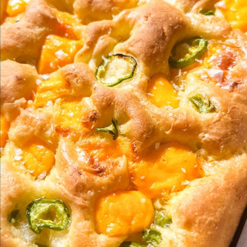 Golden focaccia bread topped with melted cheddar cheese and sliced green jalapeños, baked in a rectangular pan. The surface is slightly dimpled and crispy, with vibrant cheese and pepper accents.