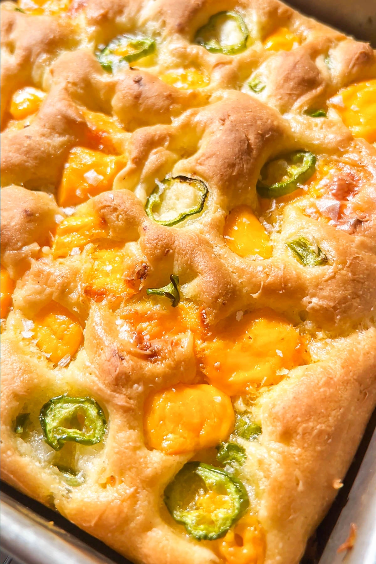 Golden focaccia bread topped with melted cheddar cheese and sliced green jalapeños, baked in a rectangular pan. The surface is slightly dimpled and crispy, with vibrant cheese and pepper accents.