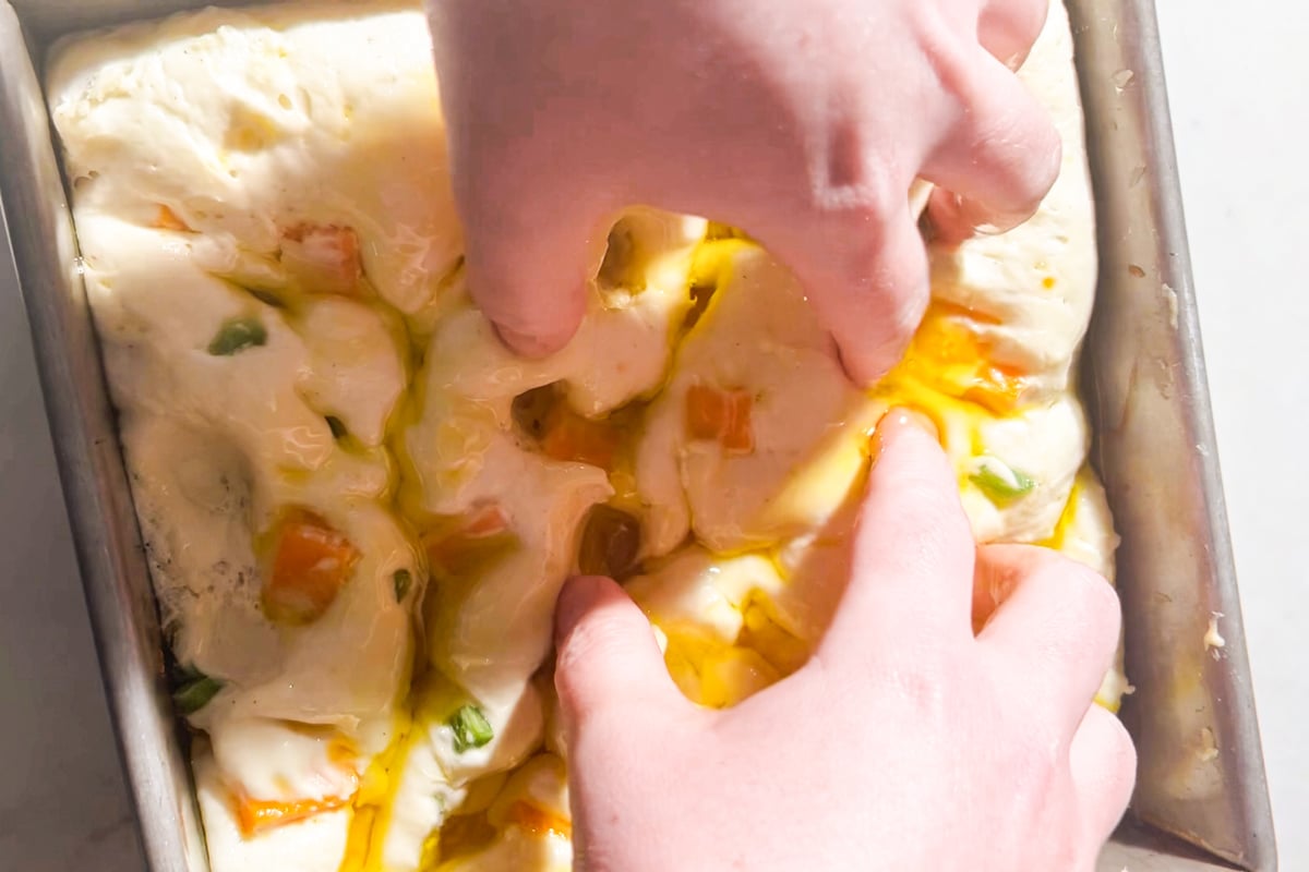Hands pressing dimples into focaccia dough in a square metal baking pan, with olive oil and small pieces of jalapeños and cheddar cheese visible on top.