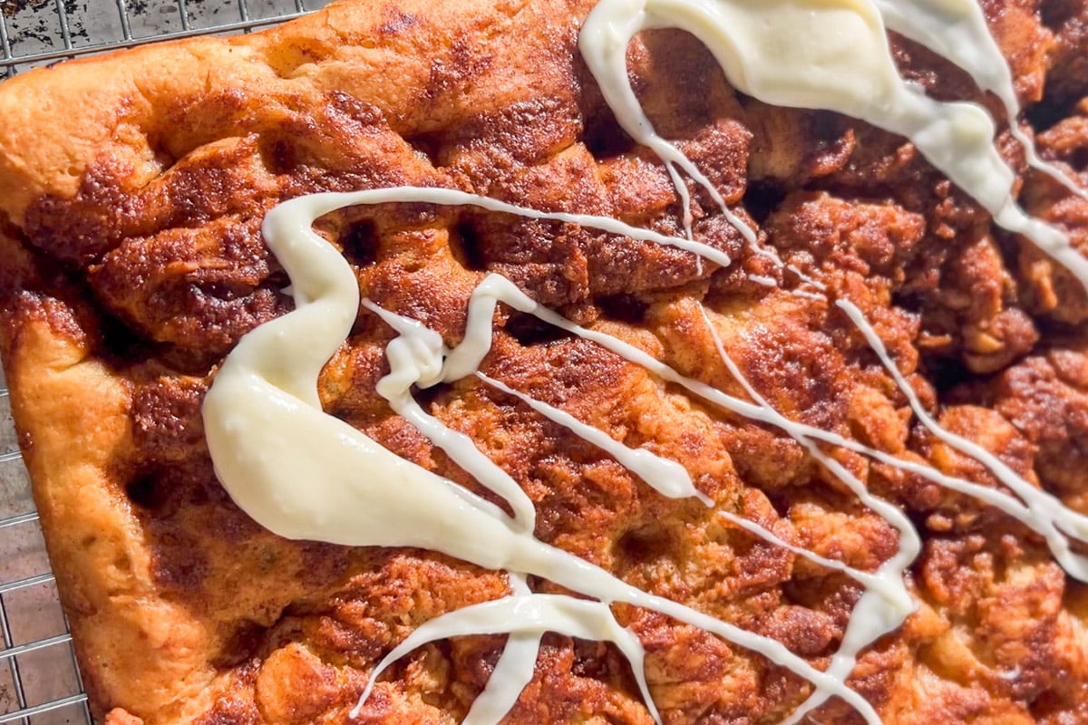 Close-up of a golden-brown cinnamon roll focaccia drizzled with cream cheese icing, resting on a wire cooling rack. The bread surface has visible dimples and swirls of cinnamon filling.