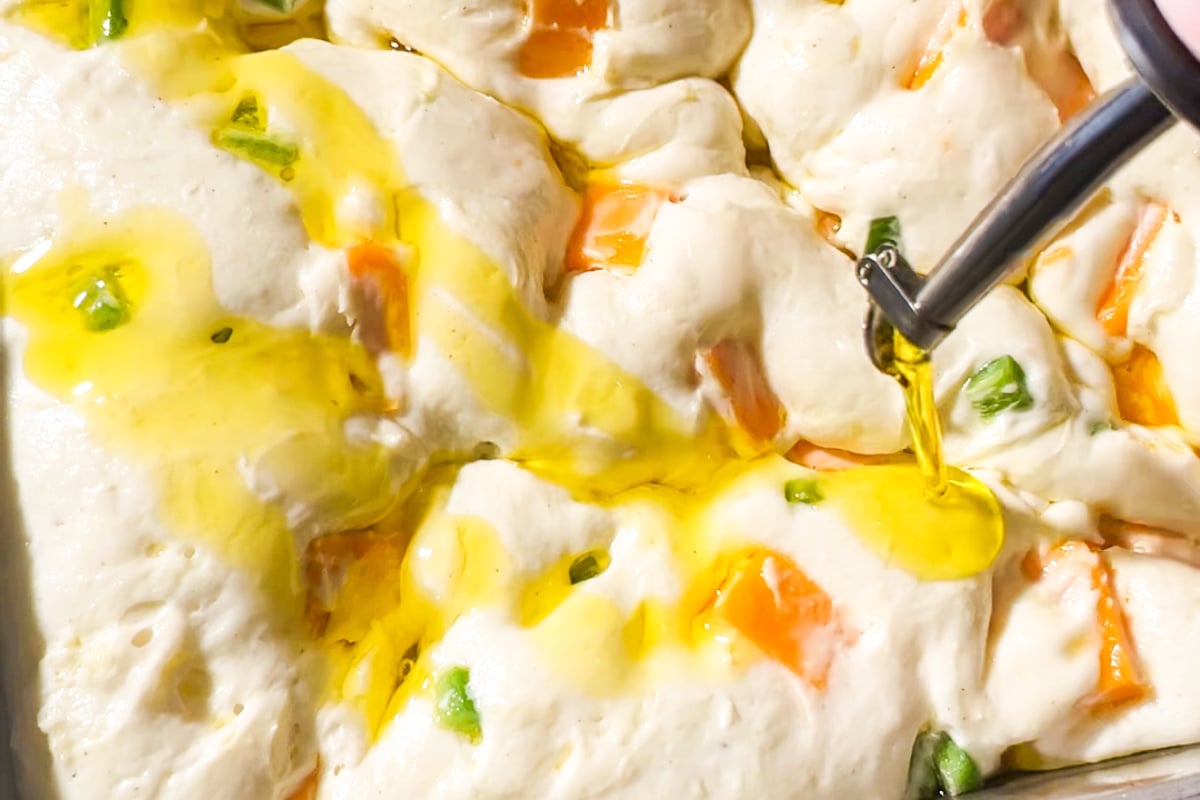 Olive oil is being poured onto unbaked gluten-free focaccia dough with visible chunks of diced jalapeño and cubed cheddar cheese in a baking pan. The dough appears soft and bubbly, ready for baking.