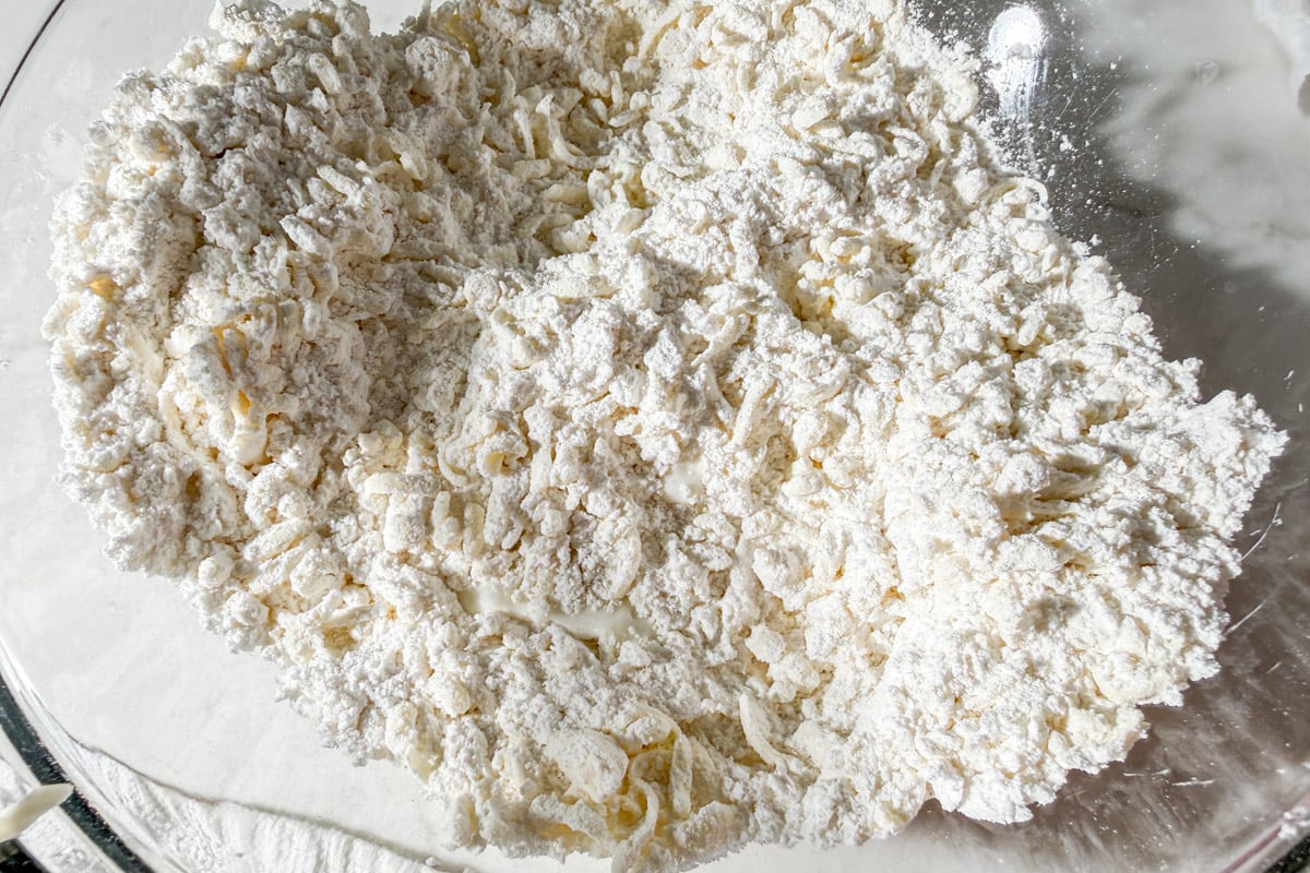 A close-up of dry ramen noodles coated in flour in a glass mixing bowl, showing a rough, powdery texture and curly noodle shapes.