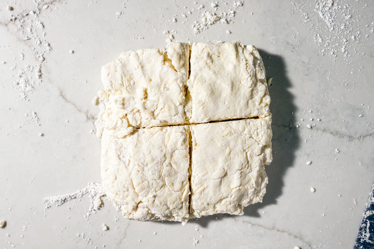 A square slab of biscuit dough on a floured marble surface, divided into four equal pieces with visible cut lines.